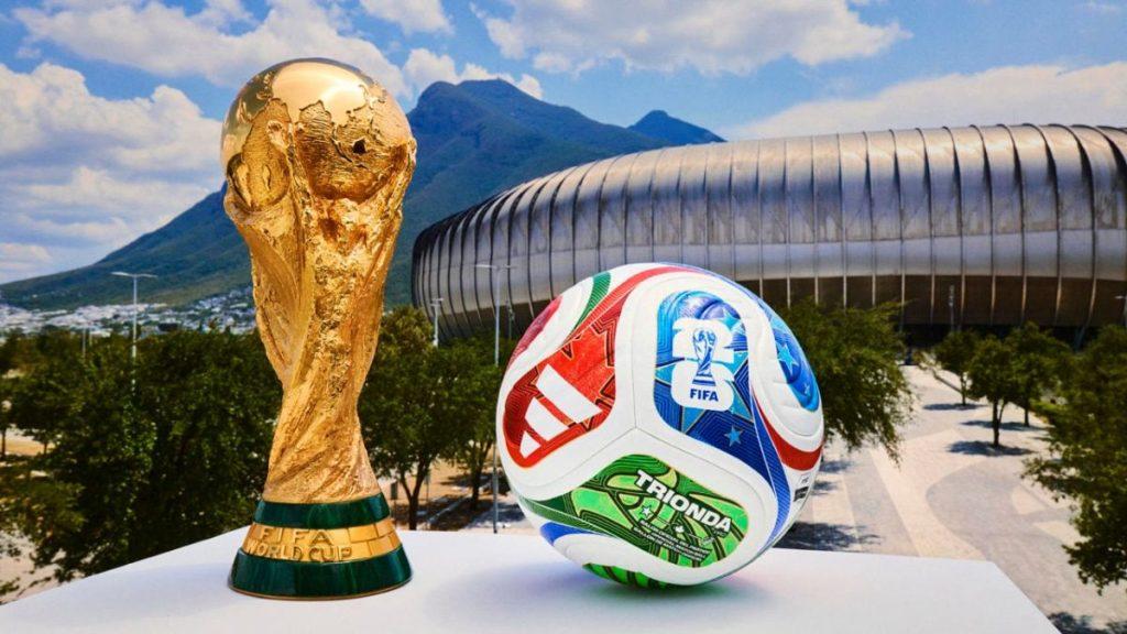Trofeo De Fifa Colocado a Lado De Un Hermoso Y Colorído Balon Del Mundial Con Un Paisaje De Montañas Y Un Estadio De Fondo.