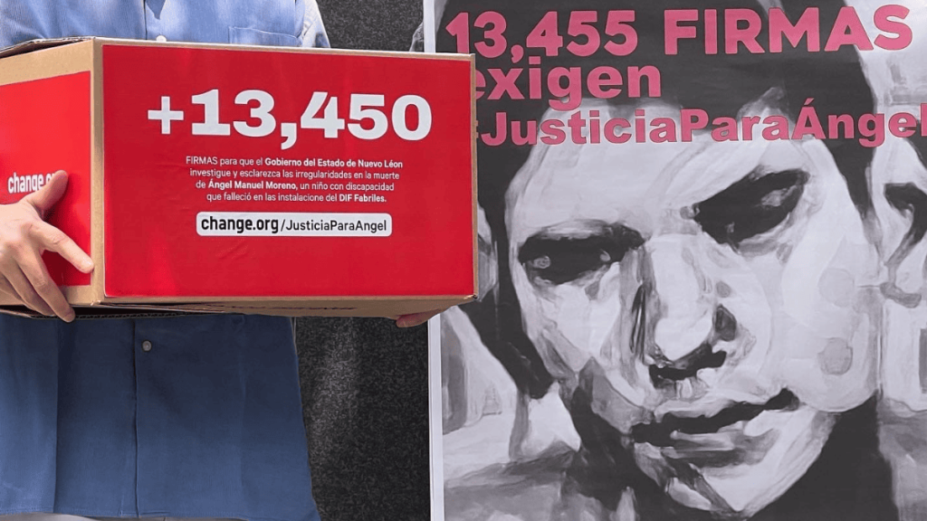 Person in a Blue Shirt Holds a Red Petition Box Showing +13,450 Signatures for Justiciaparaangel, with a Grayscale Mural in the Background. | La Silla Regia