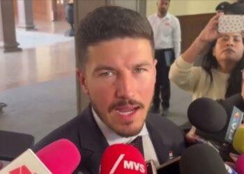 Man in a Dark Suit Speaking to Reporters, Microphones Pressed Close in a Hallway Interview Setting. | La Silla Regia