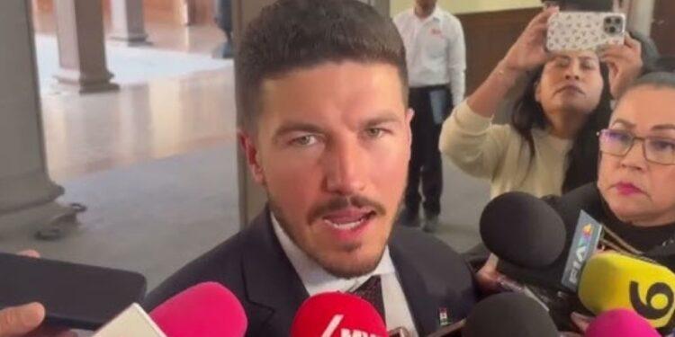 Man in a Dark Suit Speaking to Reporters, Microphones Pressed Close in a Hallway Interview Setting. | La Silla Regia