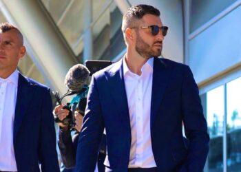 Two Men in Navy Suits Walk Outside a Glass-fronted Building, One Wearing Sunglasses While a Photographer/crew Follows with a Microphone Boom Nearby. | La Silla Regia