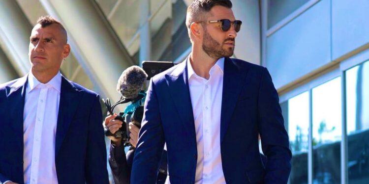 Bloque 2- Julio 5 | La Silla Regia Two Men in Navy Suits Walk Outside a Glass-fronted Building, One Wearing Sunglasses While a Photographer/crew Follows with a Microphone Boom Nearby. | La Silla Regia