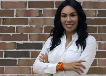 Woman with Long Dark Hair in a White Button-down Shirt, Smiling with Arms Crossed in Front of a Brick Wall. | La Silla Regia
