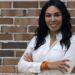 Woman with Long Dark Hair in a White Button-down Shirt, Smiling with Arms Crossed in Front of a Brick Wall. | La Silla Regia