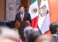 Gobernador-guerra-politica | La Silla Regia Man in a Navy Suit and Orange Tie Giving a Speech at a Podium During a Formal Event, with Mexican Flag and Another Flag in the Background. | La Silla Regia