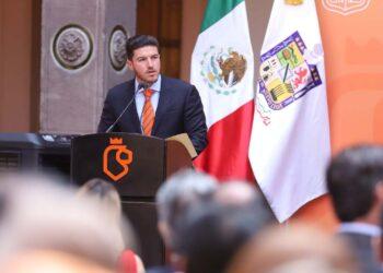 Man in a Navy Suit and Orange Tie Giving a Speech at a Podium During a Formal Event, with Mexican Flag and Another Flag in the Background. | La Silla Regia