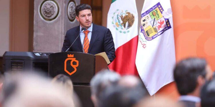 Gobernador-guerra-politica | La Silla Regia Man in a Navy Suit and Orange Tie Giving a Speech at a Podium During a Formal Event, with Mexican Flag and Another Flag in the Background. | La Silla Regia