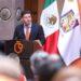 Man in a Navy Suit and Orange Tie Giving a Speech at a Podium During a Formal Event, with Mexican Flag and Another Flag in the Background. | La Silla Regia