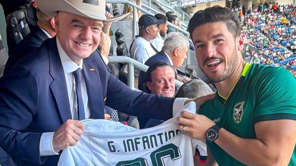 Smiling Official in a Navy Suit Hands a White Jersey to a Soccer Player in a Green Kit at a Crowded Stadium. | La Silla Regia